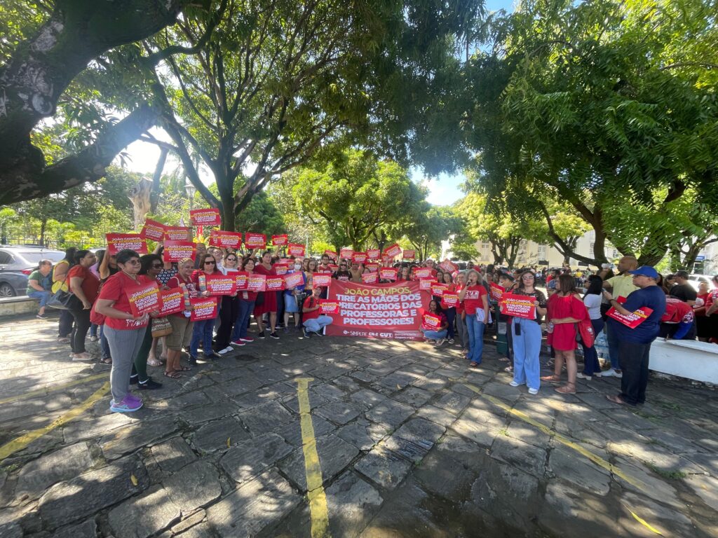 A imagem mostra uma manifestação com dezenas de professores reunidos no estacionamento da Câmara dos Vereadores do Recife. A maioria está vestindo roupas vermelhas e segura cartazes vermelhos com frases exigindo respeito aos direitos dos professores. No centro, um grande cartaz diz: “João Campos, tire as mãos dos precatórios das professoras e professores!”.