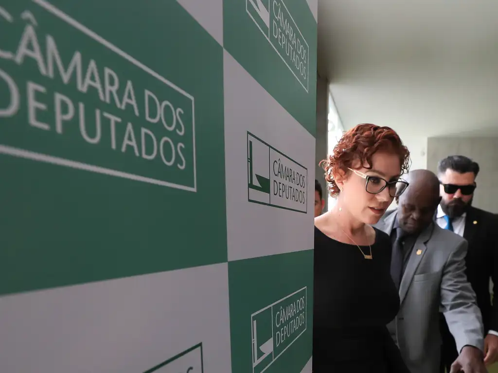 A imagem mostra a deputada federal Carla Zambelli (com cabelos curtos e cacheados, de cor ruiva, óculos de grau e vestida de preto) caminhando em um corredor da Câmara dos Deputados, em Brasília. Ela aparece ao lado de dois homens – um deles de terno cinza escuro, negro e calvo, e outro de terno preto, branco e com barba escura. Ambos usam óculos escuros, possivelmente seguranças. No lado esquerdo da imagem, há uma parede com o logotipo repetido da Câmara dos Deputados, em verde e branco. A expressão facial de Zambelli parece séria, e ela está prestes a deixar a área onde a foto foi tirada.