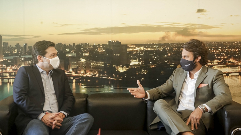 A imagem mostra o prefeito do Recife, Geraldo Julio, e Thiago Monteiro sentados em um sofá preto, conversando. Ambos usam máscaras faciais — Geraldo com máscara branca e Thiago com máscara preta com a palavra “HAUT”. Geraldo veste paletó azul e jeans; Thiago está com terno cinza claro e lenço rosa no bolso. Thiago gesticula com a mão, sugerindo que está explicando algo. Ao fundo, há uma grande imagem da cidade do Recife ao entardecer, com prédios iluminados e reflexo das luzes na água.