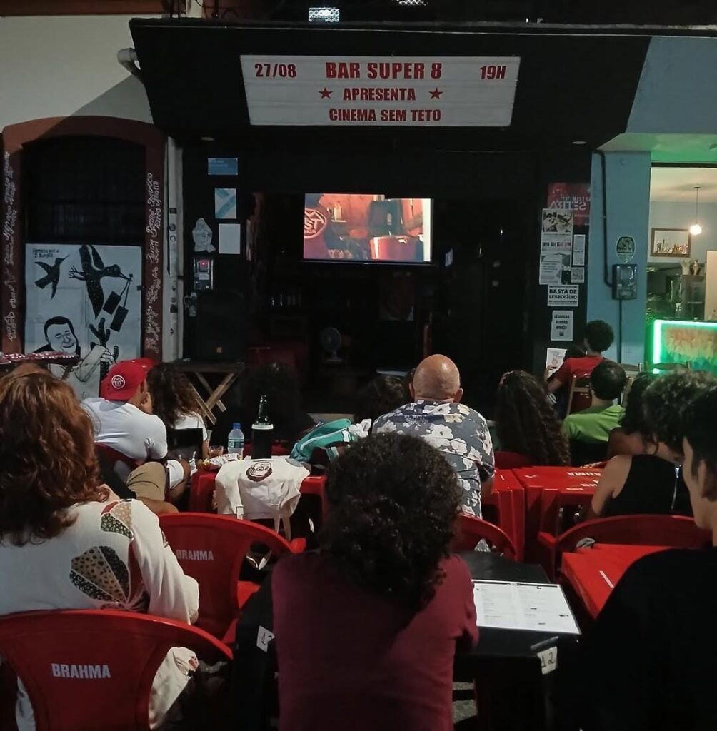 A imagem mostra um grupo de pessoas assistindo a uma sessão de cinema ao ar livre, realizada na calçada em frente a um bar chamado “Bar Super 8”. As pessoas estão sentadas em cadeiras vermelhas de plástico, muitas com a marca “Brahma” nas costas. Elas estão voltadas para uma tela que foi montada na entrada do bar, onde está sendo exibido um filme. A cena projetada mostra um ambiente interno com iluminação quente, mas não é possível identificar claramente o conteúdo. Acima da entrada do bar há uma placa que anuncia o evento: “27/08 - BAR SUPER 8 - 19H - APRESENTA - CINEMA SEM TETO”. O ambiente é informal e acolhedor, com decoração urbana e cartazes colados nas paredes. À esquerda da entrada, há um mural artístico em preto e branco com desenhos de pessoas e pássaros. Dentro do bar, vê-se uma luz verde vinda de um letreiro, sugerindo o balcão ou área de atendimento.