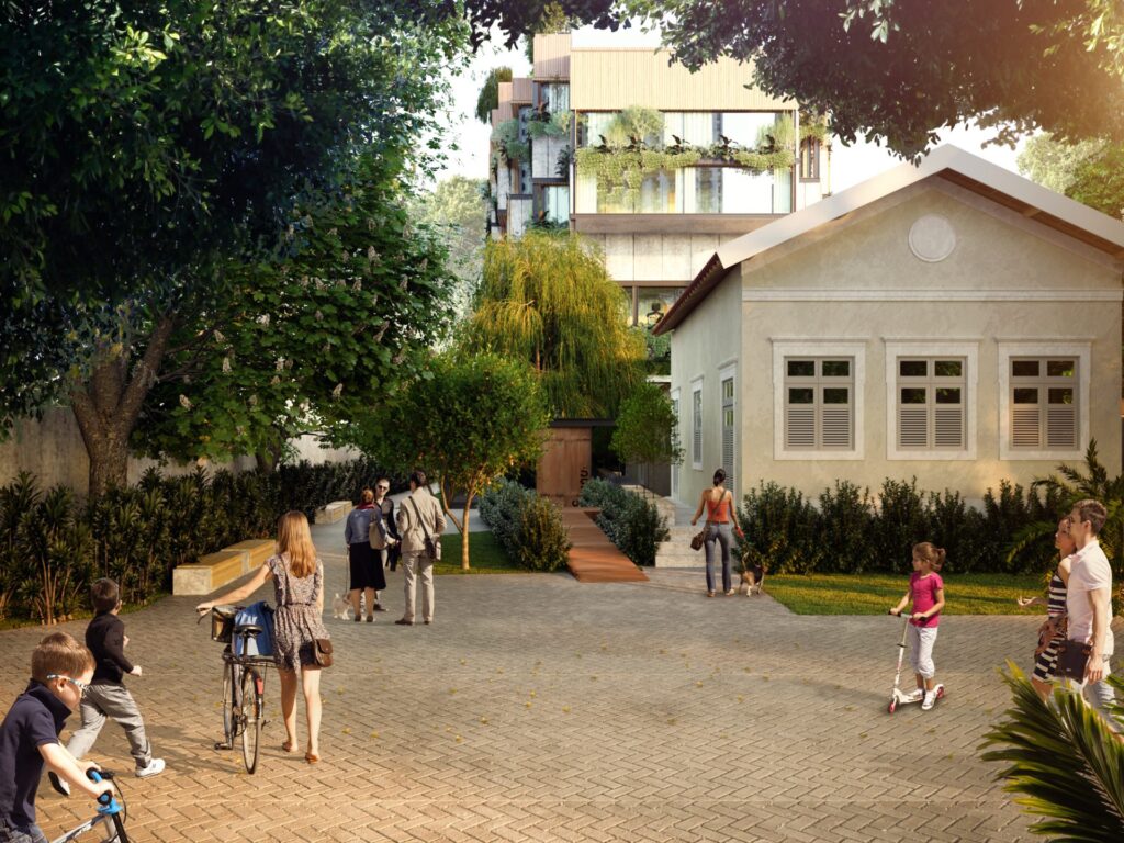 A imagem mostra um espaço urbano arborizado e bem cuidado, com pessoas caminhando, andando de bicicleta e brincando. Em primeiro plano, há um caminho pavimentado de blocos intertravados, cercado por vegetação. À esquerda, há árvores grandes e frondosas que oferecem sombra. À direita, vê-se uma casa térrea de estilo antigo, com paredes claras, janelas com venezianas brancas e um telhado de duas águas. Mais ao fundo, há um edifício moderno de vários andares com sacadas repletas de plantas, contrastando com a casa antiga. No centro da imagem, um caminho de madeira leva ao interior do terreno, onde se vê mais vegetação, incluindo uma árvore com folhas longas e pendentes. Diversas pessoas estão presentes no ambiente: uma mulher empurrando uma bicicleta, crianças brincando de bicicleta e patinete, adultos conversando, e uma mulher caminhando com um cachorro. O clima é de tranquilidade e convivência comunitária ao ar livre.