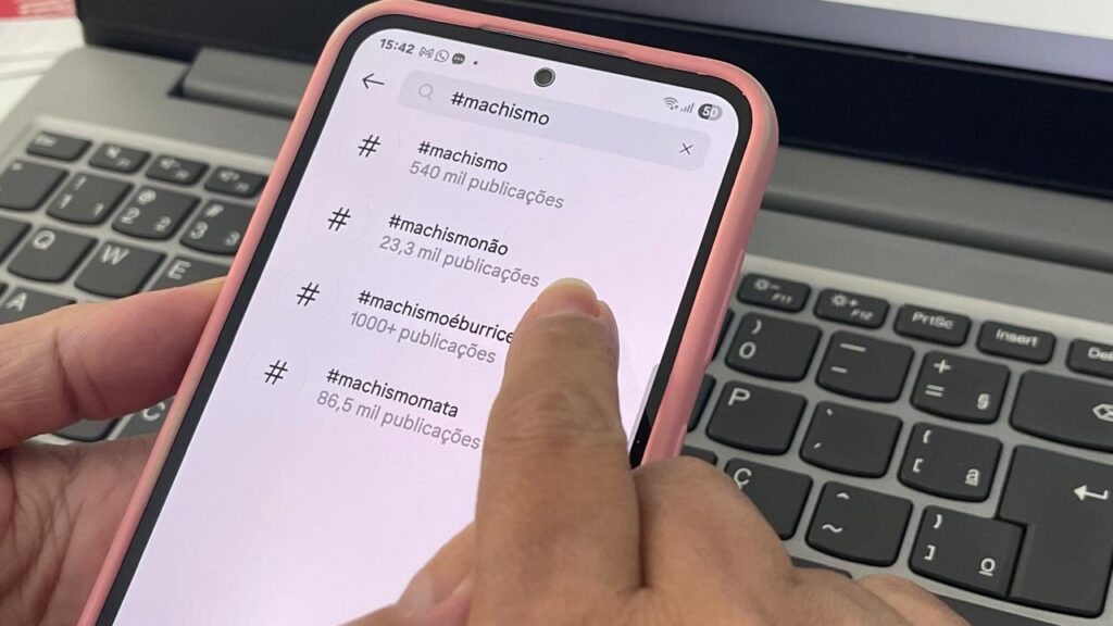 A foto mostra uma mão segurando um celular com capa rosa, em frente a um notebook com teclado visível ao fundo. Na tela do celular, está aberta uma rede social (possivelmente o Instagram), exibindo uma busca por hashtags relacionadas ao termo “machismo”. A pessoa está tocando na tela, especificamente sobre a hashtag “#machismonão”, que aparece com 23,3 mil publicações. Acima dela, está “#machismo” com 540 mil publicações. Abaixo, aparecem outras hashtags como “#machismoéburrice” com mais de mil publicações e “#machismomata” com 86,5 mil publicações.