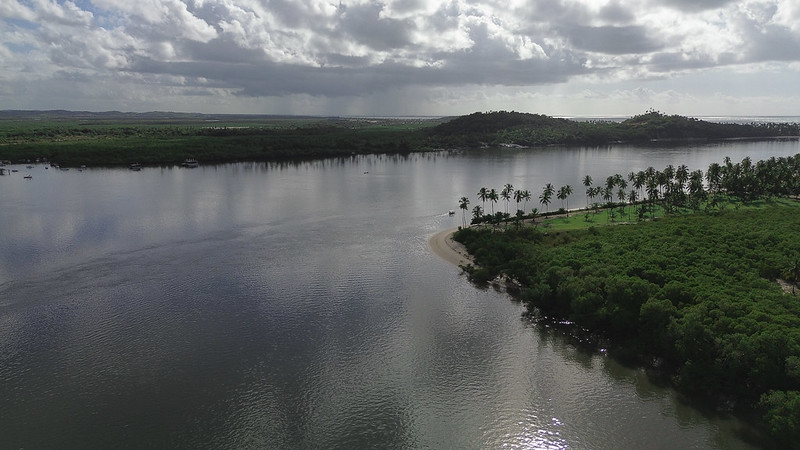 A foto, capturada de cima, mostra um rio largo e calmo que serpenteia pela paisagem natural. Em primeiro plano, à direita, há um trecho de terra coberto por vegetação verde e um grupo alinhado de coqueiros altos que margeiam uma pequena faixa de areia clara, formando uma curva suave na beira do rio. Mais adiante, do outro lado da água, há áreas extensas de manguezais e, ao fundo, morros cobertos por vegetação baixa. O céu está parcialmente nublado, com nuvens densas que projetam sombras sobre a paisagem e indicam a possibilidade de chuva ao longe, mas há também pontos de luz, onde o sol atravessa as nuvens e reflete na água.