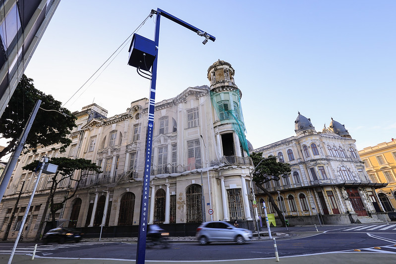 A imagem mostra uma esquina do Recife Antigo com prédios antigos e imponentes, em estilo histórico e fachada branca. O edifício principal está parcialmente coberto por andaimes e uma tela verde, indicando restauração. Na calçada, destaca-se um equipamento moderno de vigilância: uma estrutura metálica azul com uma câmera e um alto-falante no topo, usada para monitorar a rua. Alguns carros passam pela via asfaltada em frente ao prédio. O céu está claro e azul, iluminando o cenário.