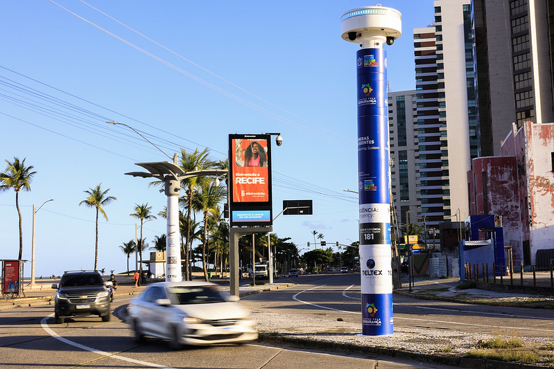 A imagem mostra uma avenida litorânea ampla e ensolarada, com alguns carros circulando. Do lado esquerdo, há coqueiros alinhados e, ao fundo, o mar e o céu azul. No centro da calçada, destaca-se um totem de segurança alto, azul e branco, equipado com câmeras e sensores no topo. Perto dele, há um painel eletrônico exibindo uma propaganda turística com a frase “Recife Te Espera”. À direita da avenida, aparecem prédios altos e modernos.