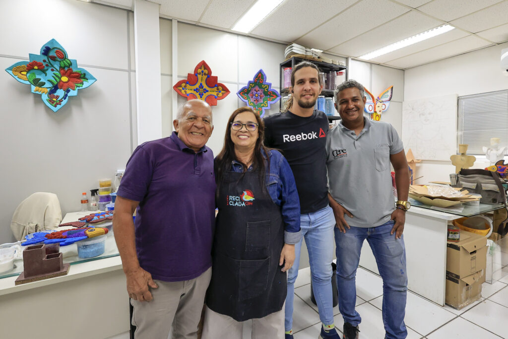 Quatro pessoas estão em pé, sorrindo para a câmera dentro de um ateliê. À esquerda, um homem idoso de camisa roxa; ao lado dele, uma mulher de avental preto, óculos e cabelos castanhos; depois, um homem jovem com camiseta preta da Reebok; e, à direita, um homem de cabelos grisalhos e camiseta cinza. Atrás deles, há mesas com materiais e enfeites coloridos em forma de mandalas na parede. O ambiente é bem iluminado, com piso branco e aparência de oficina criativa.