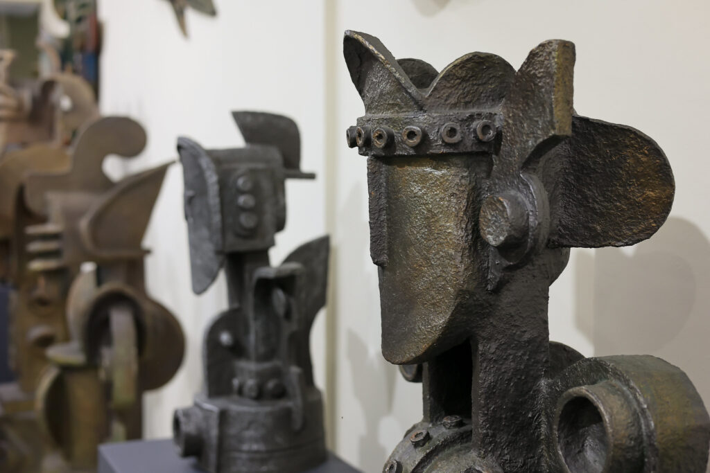 A imagem mostra uma exposição de esculturas abstratas feitas de metal ou material semelhante, com formas robustas e angulares. Em destaque, no lado direito da foto, há uma escultura que lembra um rosto estilizado com elementos que se assemelham a engrenagens ou peças industriais, como uma coroa de parafusos e orelhas em forma de discos. Ao fundo, outras esculturas com estilos similares aparecem desfocadas, alinhadas ao longo de uma parede branca. As obras têm cores escuras, com tons de ferrugem e bronze envelhecido. O ambiente tem aspecto de galeria ou museu, com iluminação clara e foco nas peças expostas.