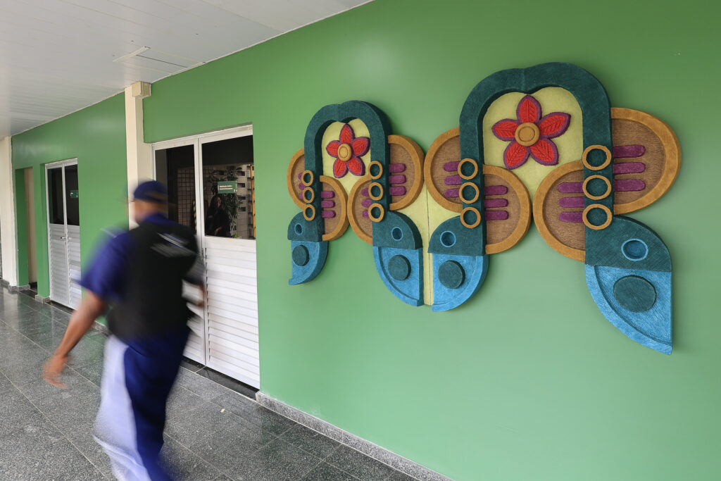 A foto mostra um corredor interno com parede verde, onde está fixada uma grande obra de arte colorida. A peça é simétrica, composta por formas curvas e circulares nas cores azul, roxo, amarelo, laranja e verde, com duas flores vermelhas ao centro. A obra lembra um par de asas estilizadas ou uma borboleta abstrata. À esquerda da imagem, um homem vestindo uniforme azul e colete preto caminha rapidamente, aparecendo em movimento borrado. Atrás dele, há portas brancas com venezianas e janelas de vidro, indicando que o local pode ser um prédio público ou escolar. O piso é escuro e brilhante, e o ambiente está bem iluminado.