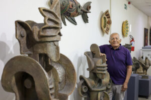 Foto de Júlio Gonçalves, um homem idoso de pele clara e cabelos brancos posa sorridente ao lado de uma grande escultura de arte em um ambiente interno, que parece ser uma galeria ou museu. Ele usa camisa polo roxa e calça clara, com uma das mãos apoiada na escultura, que tem formas geométricas e traços abstratos, lembrando um rosto estilizado. Ao redor, há outras esculturas de aparência similar, todas em tons terrosos e com aspecto robusto. A parede ao fundo exibe obras decorativas penduradas, incluindo um peixe colorido com aparência artesanal. O local é bem iluminado, com piso claro e teto de placas.