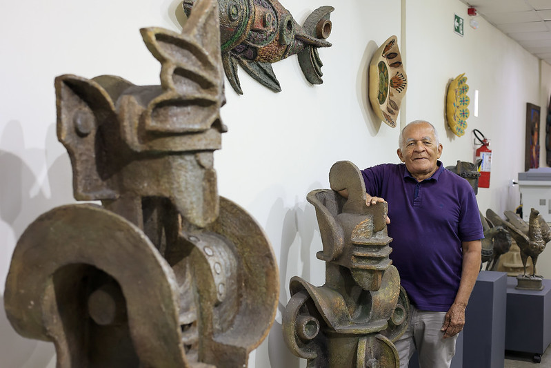 Foto de Júlio Gonçalves, um homem idoso de pele clara e cabelos brancos posa sorridente ao lado de uma grande escultura de arte em um ambiente interno, que parece ser uma galeria ou museu. Ele usa camisa polo roxa e calça clara, com uma das mãos apoiada na escultura, que tem formas geométricas e traços abstratos, lembrando um rosto estilizado. Ao redor, há outras esculturas de aparência similar, todas em tons terrosos e com aspecto robusto. A parede ao fundo exibe obras decorativas penduradas, incluindo um peixe colorido com aparência artesanal. O local é bem iluminado, com piso claro e teto de placas.