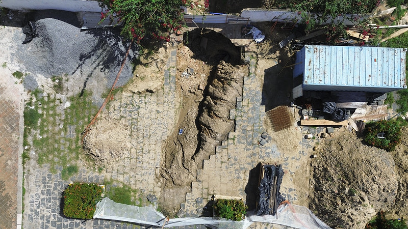 Imagem aérea mostra um sítio de escavação arqueológica em um terreno parcialmente calçado com pedras. No centro da imagem, há uma grande vala escavada no solo, com formato alongado e irregular, revelando camadas de terra. À esquerda e à direita da escavação, há montes de terra e materiais de construção. Um barracão azul-claro com telhado de zinco está localizado no canto superior direito da imagem. Há também plantas e flores ao redor do espaço, indicando que o local escavado antes era um jardim ou pátio. Alguns materiais estão cobertos por lonas e redes de proteção, sugerindo cuidados com a preservação