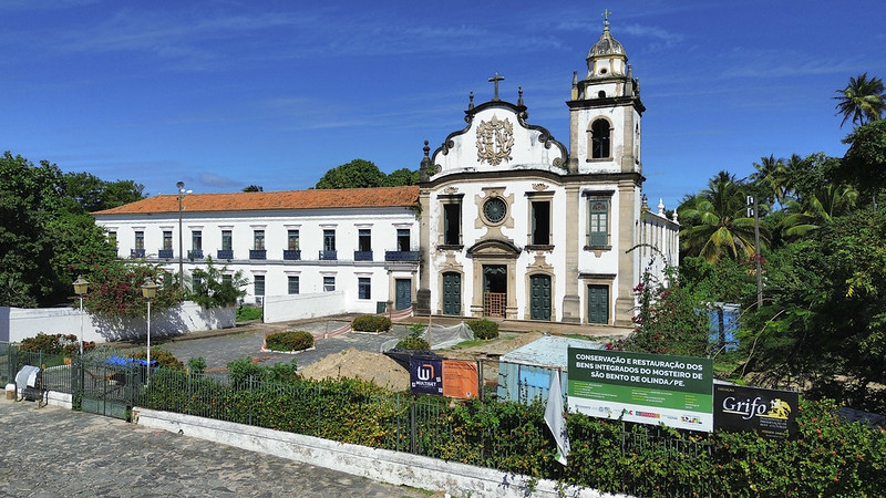 A imagem mostra a fachada do Mosteiro de São Bento de Olinda, em Pernambuco, em um dia ensolarado, com céu azul e poucas nuvens. O edifício histórico tem arquitetura colonial, com paredes brancas, detalhes em pedra escura e um telhado de barro. No centro, destaca-se a igreja com uma torre à direita e uma fachada ricamente ornamentada, incluindo uma grande cruz no topo. À frente do mosteiro, há uma área cercada onde ocorrem escavações arqueológicas. No canteiro, vê-se um contêiner azul, lonas, placas informativas e banners com logotipos de instituições ligadas ao projeto de pesquisa e preservação. Ao redor, há árvores e vegetação típica do clima tropical.