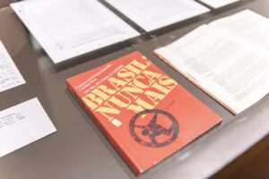 Fotografia em ambiente de exposição. No centro da imagem, sobre uma superfície plana, está o livro Brasil: Nunca Mais, com a capa predominantemente vermelha e o título em letras grandes e amarelas. Abaixo do título, há uma ilustração preta de uma figura humana presa dentro de uma engrenagem, simbolizando repressão. Acima do título, lê-se: “Projeto da Pastoral Evangélica - Um relato para a história”. O exemplar exposto é da 17ª edição. Ao redor do livro, há documentos datilografados e páginas abertas de outras publicações, todos protegidos sob uma vitrine de vidro. À esquerda, um pequeno cartão exibe informações bibliográficas da obra. A iluminação é clara, destacando o material exposto.