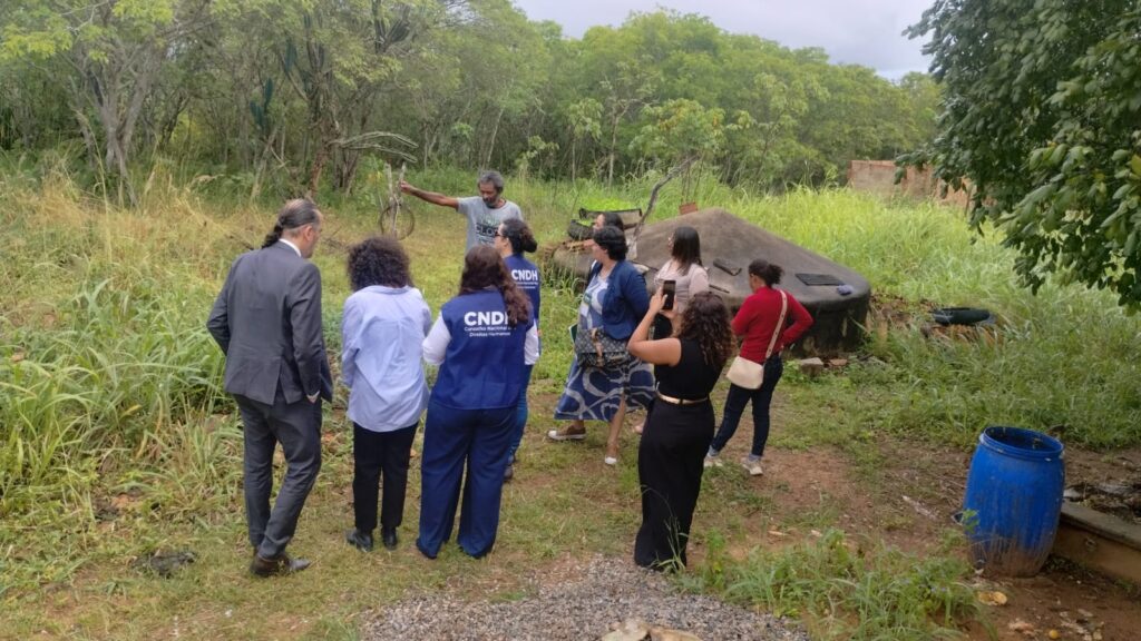 A fotografia mostra um grupo de cerca de dez pessoas reunidas em uma área de vegetação aberta, com grama alta, árvores e arbustos ao fundo. À esquerda da imagem, um homem com cabelos grisalhos e barba, vestindo camiseta cinza, está de pé e gesticula com o braço estendido, como se explicasse algo ao grupo. As demais pessoas estão viradas para ele, ouvindo atentamente. Entre elas, há mulheres e homens com diferentes estilos de roupa — alguns mais formais, outros casuais. Uma das mulheres veste um colete azul com a sigla CNDH, que significa Conselho Nacional de Direitos Humanos. À direita da cena, há uma estrutura arredondada de concreto parcialmente coberta de vegetação, possivelmente uma cisterna ou fossa séptica, e também um barril azul de plástico. O ambiente transmite a sensação de uma visita de campo ou inspeção em área rural.