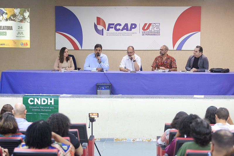 A foto mostra uma mesa de debates com cinco pessoas sentadas, em um auditório da Universidade de Pernambuco (UPE), na Faculdade de Administração e Direito (FCAP). Há um banner ao fundo com os logotipos da UPE e da FCAP, além de um cartaz anunciando uma atividade da Relatoria Nacional em Pernambuco. Na mesa, há homens e uma mulher, todos com microfones e materiais de apoio. Um dos participantes fala enquanto os demais escutam. Em frente à mesa, há uma faixa verde com o nome do Conselho Nacional dos Direitos Humanos (CNDH) e pessoas assistem ao evento na plateia.