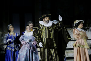Em um palco ao ar livre, seis pessoas vestem trajes de época inspirados no período barroco europeu, lembrando o século XVII. O foco está em um homem no centro da imagem, que usa um elaborado traje preto com listras douradas, gola branca volumosa e chapéu de abas largas com enfeite dourado. Ele tem barba, expressão séria e aponta um dedo para o alto, em gesto teatral. Ao redor dele, outras pessoas — homens e mulheres — usam vestidos longos e coloridos, com babados, rendas e adornos típicos da nobreza da época. Todos parecem estar envolvidos em uma encenação histórica ou apresentação cultural. O fundo está escuro, destacando os figurinos iluminados.