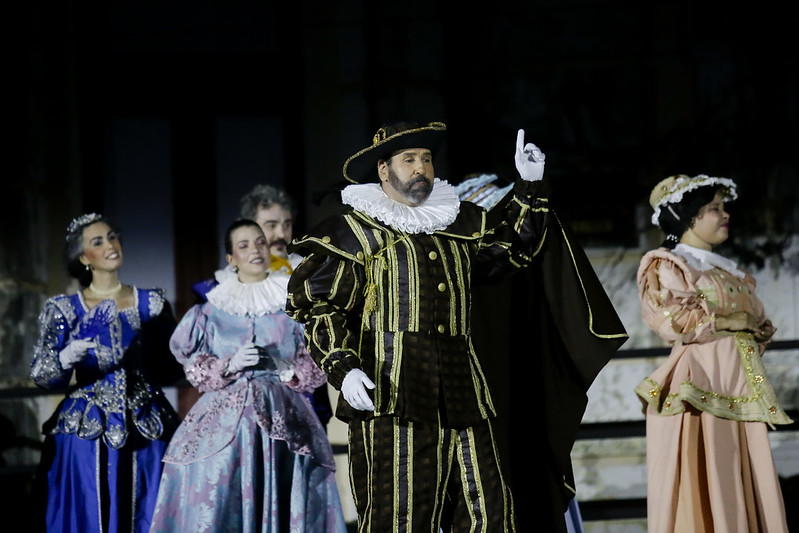 Em um palco ao ar livre, seis pessoas vestem trajes de época inspirados no período barroco europeu, lembrando o século XVII. O foco está em um homem no centro da imagem, que usa um elaborado traje preto com listras douradas, gola branca volumosa e chapéu de abas largas com enfeite dourado. Ele tem barba, expressão séria e aponta um dedo para o alto, em gesto teatral. Ao redor dele, outras pessoas — homens e mulheres — usam vestidos longos e coloridos, com babados, rendas e adornos típicos da nobreza da época. Todos parecem estar envolvidos em uma encenação histórica ou apresentação cultural. O fundo está escuro, destacando os figurinos iluminados.