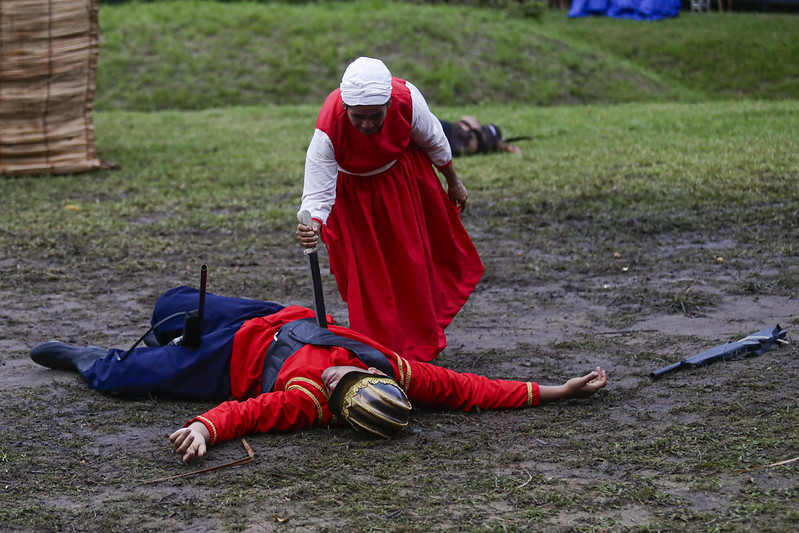 Em uma encenação teatral ao ar livre, uma mulher de vestido longo vermelho e touca branca se inclina sobre um homem caído no chão, como se estivesse cravando uma espada em seu corpo. O homem veste uniforme vermelho com calça azul e capacete dourado, e está estirado de costas na lama, com os braços abertos. Ao fundo, sobre a grama, outro corpo simula estar morto, reforçando o clima de batalha. A cena ocorre em um espaço gramado, com um escudo de palha à esquerda e vegetação ao redor, sugerindo uma representação histórica de combate.