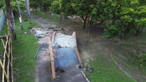A imagem mostra uma área de parque com muitas árvores e um caminho pavimentado que passa por obras. No centro da imagem, há uma ponte de concreto danificada, com a parte superior quebrada e coberta por entulhos. Os parapeitos laterais de tijolos estão rachados e tombados. Ao redor da ponte, há estruturas metálicas azuis de proteção, algumas caídas no chão. A vegetação ao redor é abundante, com grama e árvores de médio porte. À esquerda da imagem, vê-se um cercado de madeira e uma cerca de tela azul. Um poste de iluminação também aparece, inclinado, próximo ao trecho em obras.