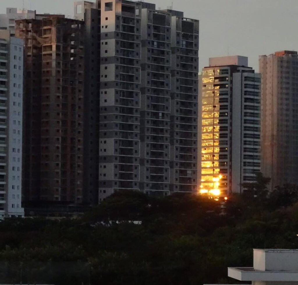 Esta é uma fotografia urbana feita durante o entardecer. A imagem mostra vários prédios altos, a maioria com fachadas escuras ou em tons acinzentados, dois deles ainda em construção ou inacabados. No entanto, entre essas construções escuras, há um prédio de fachada espelhada que se destaca intensamente. Esse edifício reflete diretamente os raios do sol poente, criando uma faixa vertical dourada e brilhante que parece uma coluna de fogo ou uma fenda luminosa entre os prédios sombreados. A luz é tão intensa que dá a impressão de que o próprio prédio está emitindo luz, como se estivesse aceso por dentro com uma chama dourada. Na parte inferior da imagem, há uma área com muitas árvores escuras, provavelmente um parque ou conjunto de copas, contrastando com o céu claro e os edifícios altos ao fundo.