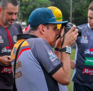 A foto mostra um homem chamado Teco Barbero, que é deficiente visual, fotografando com uma câmera profissional em um ambiente ao ar livre, provavelmente um campo de futebol. Ele está de perfil, com o olho encostado no visor da câmera, em plena ação de registrar uma imagem. Teco veste uma camisa esportiva com as palavras “Comissão Técnica” nas costas e logos da Prefeitura de Sorocaba e outros patrocinadores. Ele usa boné azul, relógio digital e carrega a câmera presa por uma alça no pescoço. Ao fundo, outros homens também vestem uniformes semelhantes e parecem estar envolvidos em um evento esportivo. A imagem transmite concentração e quebra de estereótipos, mostrando uma pessoa com deficiência visual atuando como fotógrafo profissional.