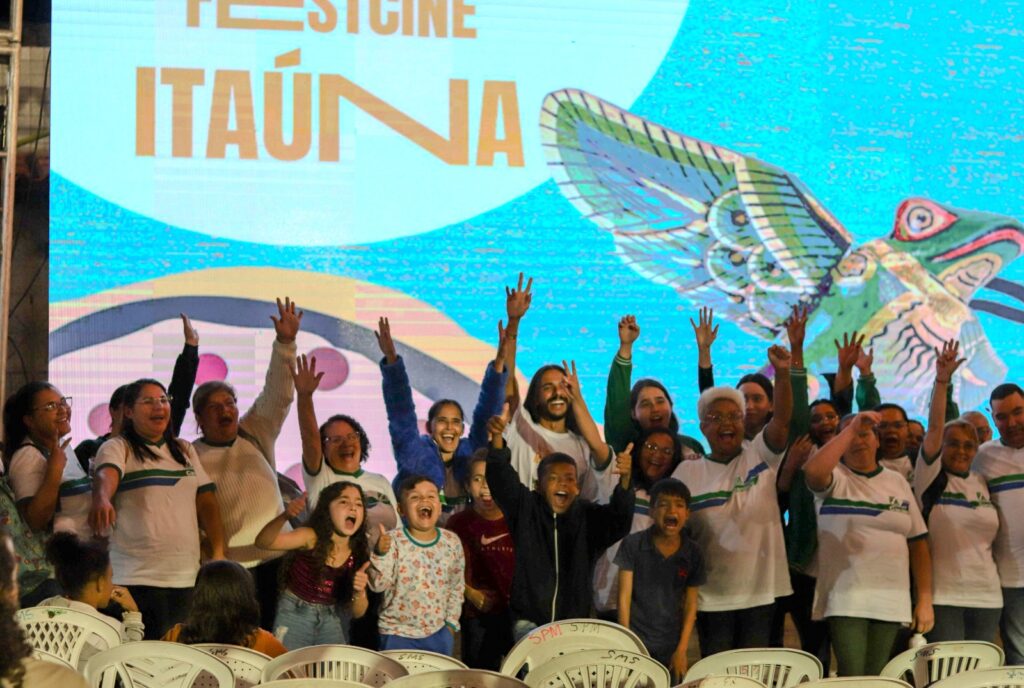 A foto mostra um grupo de cerca de 20 pessoas reunidas em um palco, comemorando de forma animada. Elas estão com os braços erguidos e sorrindo, aparentando felicidade. Entre elas, há crianças, adolescentes e adultos, a maioria usando camisetas brancas com uma faixa azul e verde no peito. Atrás do grupo, há um grande telão colorido com a imagem de um pássaro estilizado, que parece um beija-flor, e os dizeres “FESTCINE ITAÚNA”. Na frente do palco, há várias cadeiras plásticas brancas voltadas para o público. A cena transmite um clima de celebração, possivelmente relacionado a um festival de cinema.