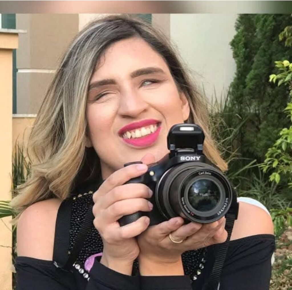 Na imagem, Micheli Paula de Oliveira aparece sorridente segurando uma câmera profissional da marca Sony. Ela veste uma blusa preta de alças com recortes nos ombros e detalhes brilhantes. Seu cabelo loiro com mechas está solto, levemente ondulado. Micheli está ao ar livre, diante de um jardim com folhagens verdes e um prédio ao fundo. Seu olhar está voltado levemente para cima, com expressão de alegria e realização. A foto transmite leveza, empoderamento e a essência da sua trajetória como cinegrafista com deficiência visual.