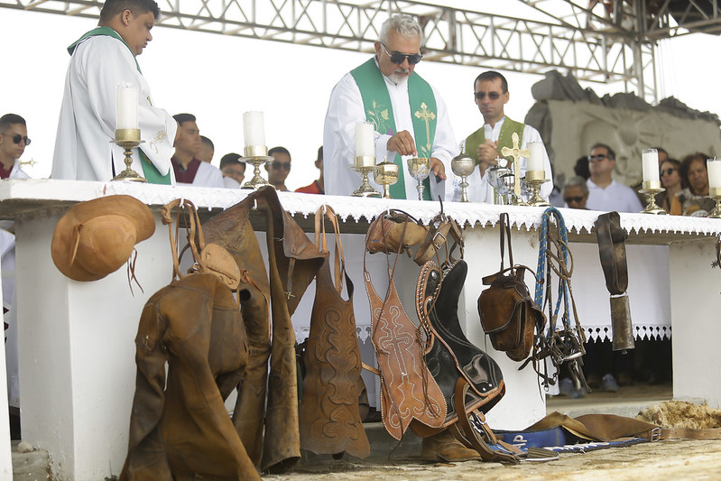A imagem mostra uma missa ao ar livre celebrada por padres vestidos com paramentos verdes e brancos. Eles estão atrás de um altar coberto com uma toalha branca e enfeitado com castiçais, cálices e objetos litúrgicos. Na parte da frente do altar, estão pendurados diversos itens do vestuário tradicional de vaqueiros nordestinos: gibões de couro, perneiras, chapéus, selas e outros acessórios usados na lida com o gado. Ao fundo, várias pessoas assistem à cerimônia com atenção.