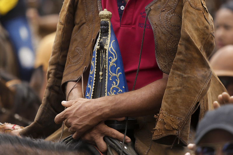 Foto mostra o tronco de um homem montado a cavalo, segurando com firmeza uma pequena imagem de Nossa Senhora Aparecida, a padroeira do Brasil. A santa tem manto azul escuro decorado com detalhes dourados e coroa na cabeça. O homem veste camisa vermelha e um gibão de couro marrom — vestimenta tradicional dos vaqueiros do Nordeste. Ao fundo, fora de foco, há outras pessoas e animais, sugerindo um ambiente de romaria ou celebração popular.