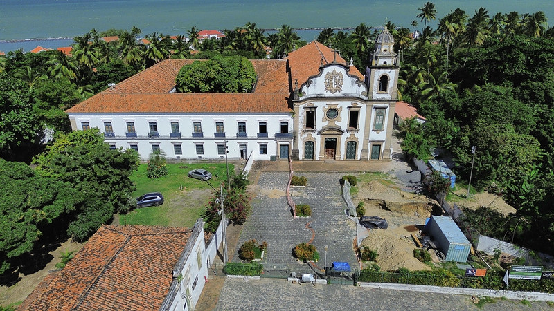 A imagem aérea mostra o Mosteiro de São Bento de Olinda, em Pernambuco, visto de frente e ao alto. O conjunto arquitetônico tem estilo colonial, com paredes brancas e telhado de telhas cerâmicas. À esquerda está o edifício do mosteiro, longo e retangular, e ao centro a igreja, com fachada ornamentada e uma torre com sino à direita. Em frente ao prédio, há um pátio de pedra com canteiros e uma área em obras: escavações arqueológicas estão em andamento, com valas abertas, montes de terra e um contêiner azul usado como apoio. Ao redor, há vegetação abundante, incluindo coqueiros e árvores frondosas. Ao fundo, vê-se o mar e parte da costa urbana de Olinda, compondo uma paisagem histórica e natural integrada.