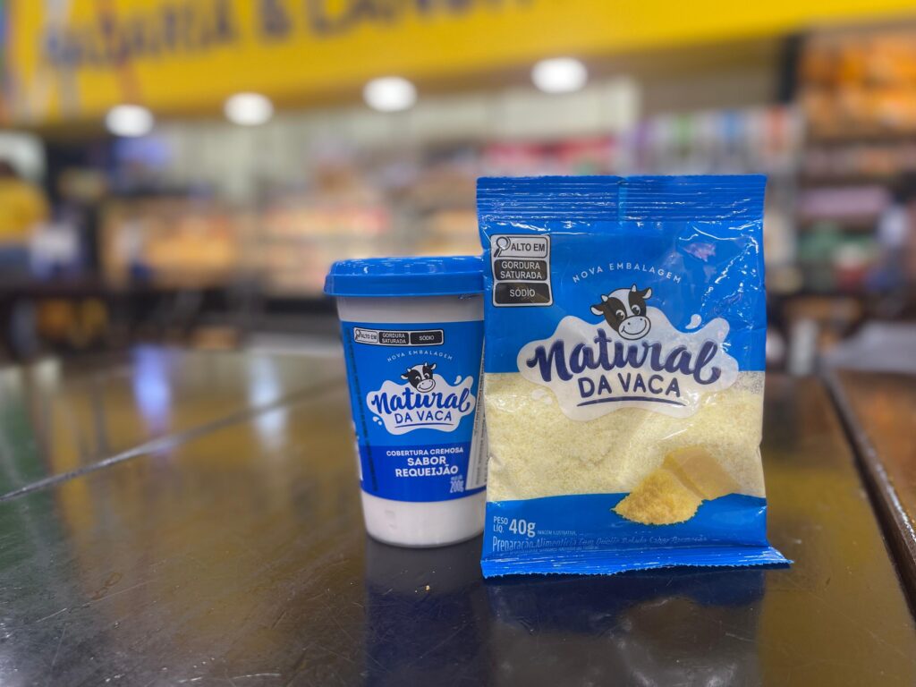 A foto mostra dois produtos da marca Natural da Vaca, expostos sobre uma superfície metálica brilhante em um supermercado. À esquerda, há um pote azul e branco com tampa azul, contendo cobertura cremosa sabor requeijão, com 200 gramas. À direita, está um pacote azul e transparente com queijo ralado de cor amarela clara, pesando 40 gramas. Ambos os produtos exibem o logotipo de uma vaca sorridente. Na embalagem do queijo, há alertas de alto teor de gordura saturada e sódio. O fundo da imagem está desfocado, mas revela prateleiras e uma parede amarela com letras grandes. A iluminação do ambiente é clara e suave. A composição destaca os produtos em primeiro plano.