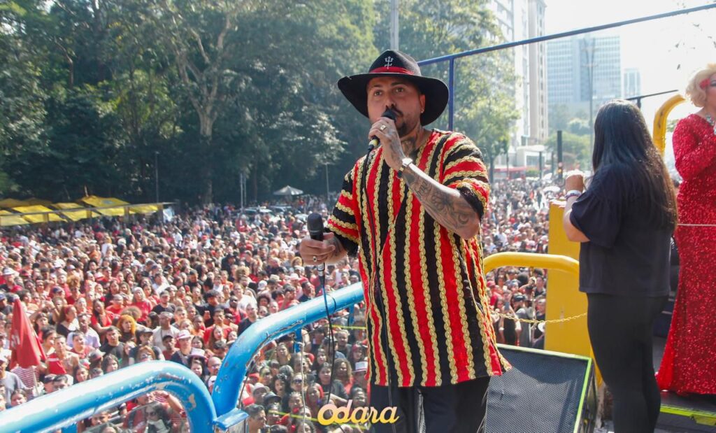 A foto mostra Jonathan Pires cantando em cima de um trio elétrico diante de uma grande multidão. Ele segura um microfone na mão direita e veste um chapéu preto de abas largas e uma camisa listrada nas cores preta, vermelha e amarela. Tem tatuagens visíveis nos braços e usa relógio no pulso. Ao fundo, vê-se uma imensa plateia reunida na rua, muitas pessoas vestidas de vermelho e preto, transmitindo clima de festa popular. Atrás dele, outras pessoas também estão no trio, incluindo uma artista com vestido vermelho brilhante. O cenário é de evento ao ar livre, com árvores altas de um lado e prédios da cidade ao fundo.