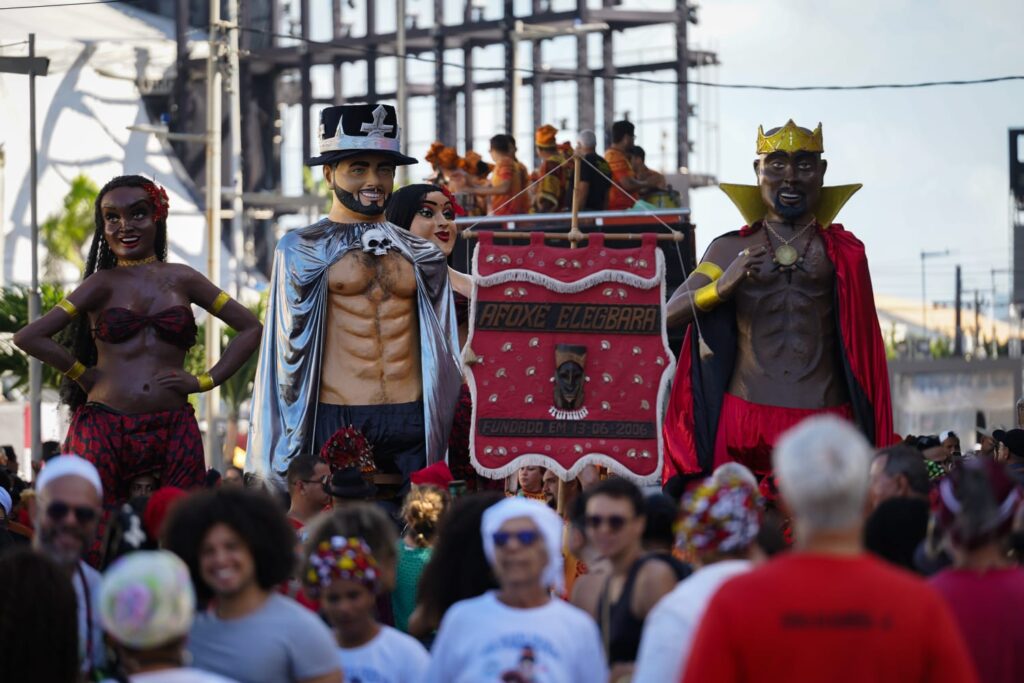 A foto mostra um cortejo de carnaval nas ruas, com um grupo de pessoas desfilando ao redor de grandes bonecos típicos do carnaval de Pernambuco, conhecidos como bonecos gigantes. No centro, há um estandarte vermelho com o nome “Afoxé Elegbára” e a data de fundação “13-06-2006”. Atrás do estandarte, aparecem três bonecos gigantes: à esquerda, uma mulher negra com roupa vermelha estampada e acessórios dourados; no meio, um homem de chapéu preto e capa prateada, exibindo o peito nu e musculoso; e à direita, outro homem negro com coroa dourada, capa vermelha e colar com medalhão. À frente, muitas pessoas acompanham o cortejo, algumas usando roupas brancas e turbantes, outras com camisas coloridas e cabelos soltos.