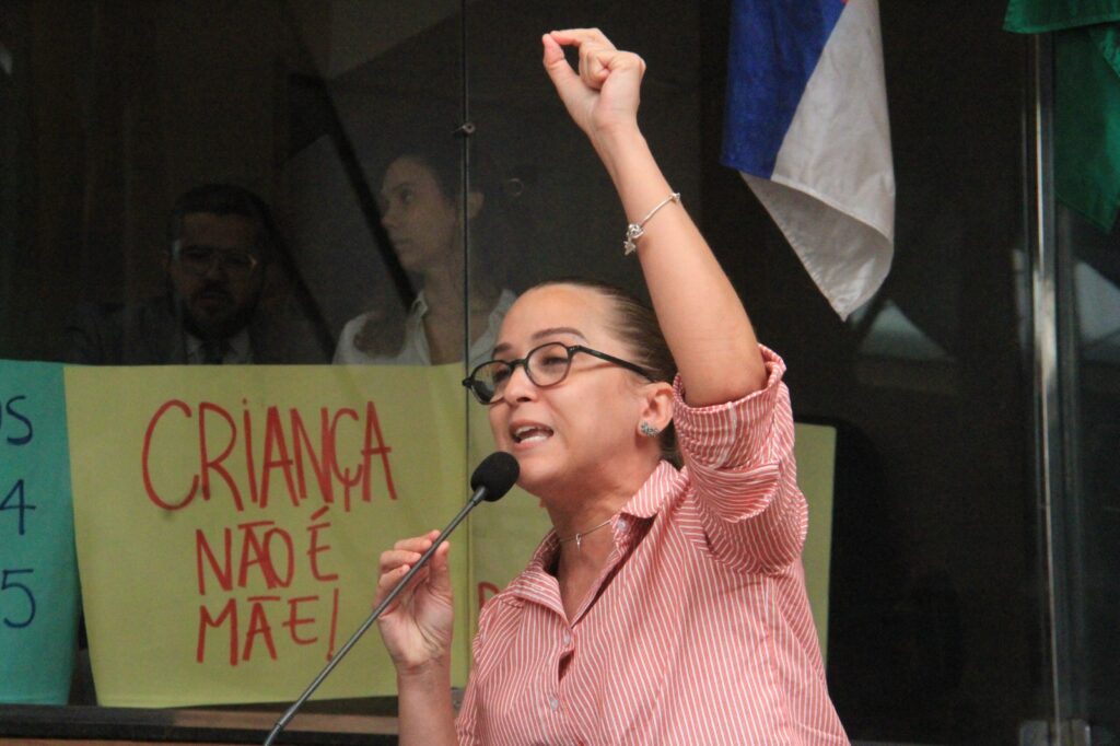 A foto mostra a vereadora Kari Santos discursando em um ambiente que parece ser uma audiência pública ou sessão oficial. Ela fala ao microfone com expressão firme e braço erguido em gesto de determinação. Usa óculos de armação escura, tem cabelo preso e veste uma blusa avermelhada com riscas brancas. Atrás dela, um cartaz manuscrito em letras vermelhas traz a frase “CRIANÇA NÃO É MÃE!”, mensagem de protesto clara e incisiva. O ambiente é formal, com outras pessoas desfocadas ao fundo. O tom geral da cena transmite engajamento político e luta por direitos, destacando a energia e a convicção da vereadora.