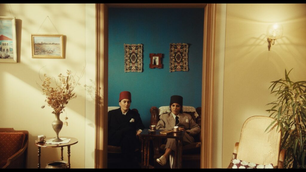 A imagem mostra uma sala de estar com decoração clássica e elegante. Duas mulheres vestidas com ternos e chapéus estão sentadas em poltronas no centro. À esquerda, há uma mesa com flores secas e quadros na parede. Ao fundo, a parede azul exibe tapetes pendurados e uma foto central. À direita, vê-se uma cadeira de vime e uma planta alta.