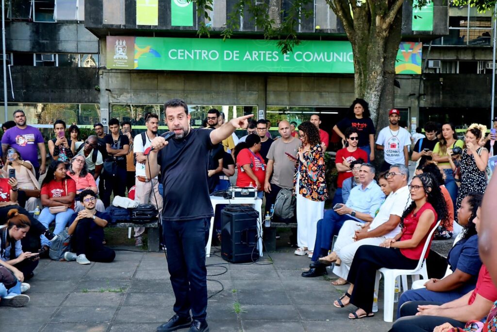 De camisa preta, Guilherme Boulos fala ao microfone e gesticula enquanto se dirige a um grupo numeroso de pessoas reunidas em frente ao Centro de Artes e Comunicação da UFPE. Parte do público está sentada, outra em pé, e alguns registram o momento com celulares.