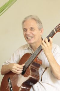 A imagem mostra um homem, com cabelos e pele branca, sorrindo enquanto toca um violão marrom.