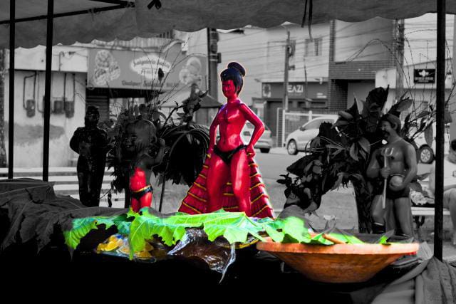 A imagem mostra exposição de estatuetas em uma mesa ao ar livre. No centro, destaca-se uma figura de Exú destacada em vermelho, cercada por outras esculturas e elementos naturais como folhas e frutas. O contraste entre as cores vivas e o fundo em preto e branco realça a força cultural da cena, representando a arte popular e a ancestralidade.