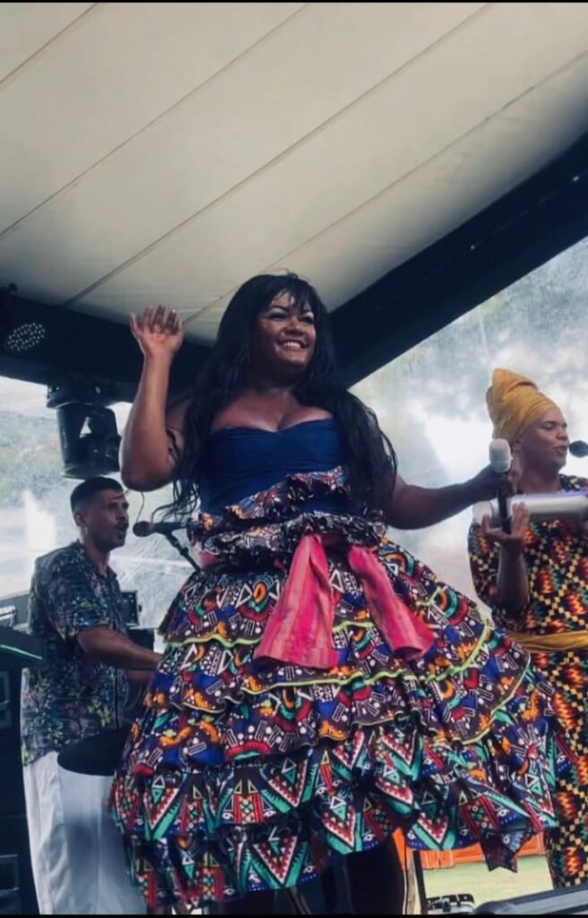 A foto mostra Raphaella Ribeiro, uma mulher negra sorridente em pé, com uma pose animada. Ela veste um top azul escuro e uma saia ampla com padronagem colorida vibrante, adornada com um laço rosa na frente. Ao fundo, outras pessoas e um teto claro sugerem um ambiente interno, possivelmente uma apresentação ou encontro festivo.