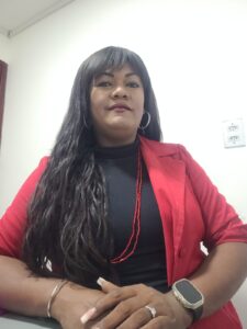 A foto mostra Raphaella Ribeiro uma mulher de pele em tom médio, cabelos longos e escuros com franja, vestindo blazer vermelho sobre blusa preta, colar de contas vermelhas e brincos de argola. Ela está sentada em ambiente interno, diante de uma parede branca com um interruptor ao fundo, e apoia as mãos suavemente à frente. Sua expressão é serena e confiante, com leve sorriso e postura elegante.