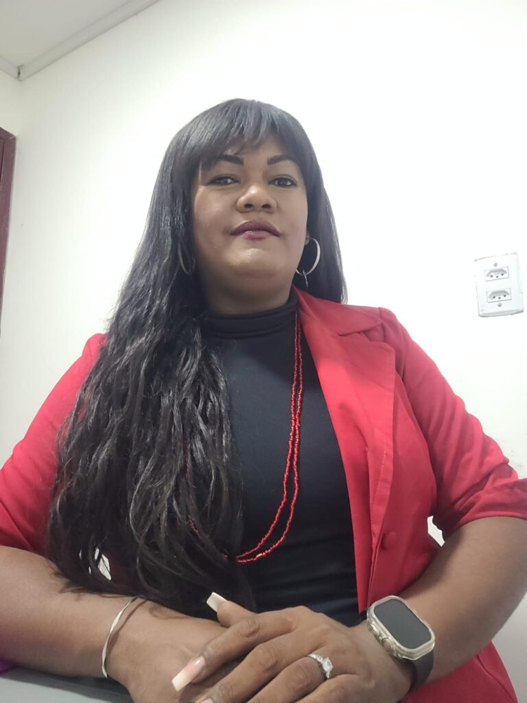 A foto mostra Raphaella Ribeiro uma mulher de pele em tom médio, cabelos longos e escuros com franja, vestindo blazer vermelho sobre blusa preta, colar de contas vermelhas e brincos de argola. Ela está sentada em ambiente interno, diante de uma parede branca com um interruptor ao fundo, e apoia as mãos suavemente à frente. Sua expressão é serena e confiante, com leve sorriso e postura elegante.