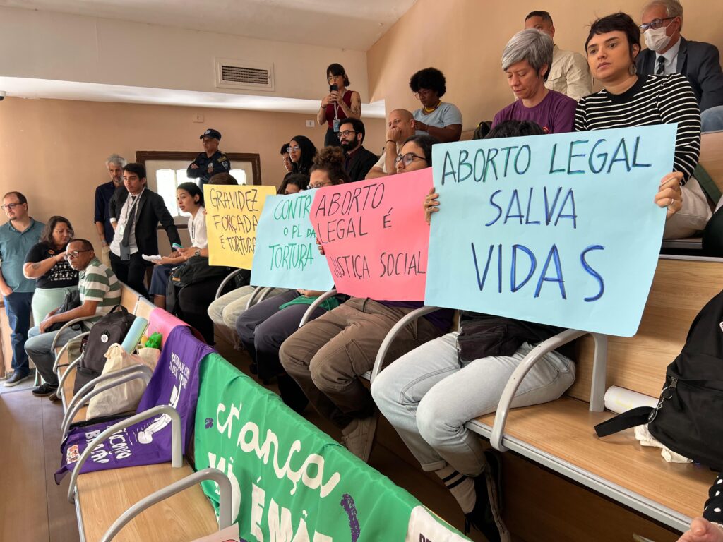 A foto mostra uma sala ampla, parecida com um plenário ou auditório público, onde várias pessoas acompanham uma sessão. Em primeiro plano, três mulheres estão sentadas lado a lado, segurando cartazes coloridos feitos à mão. As mensagens, escritas em letras grandes, expressam apoio à legalização do aborto e denunciam a gravidez forçada como forma de violência. O tom geral dos cartazes é de protesto e reivindicação de direitos, com frases curtas, diretas e impactantes. As cores vibrantes — como rosa, azul, verde e amarelo — reforçam a visibilidade e a força do ato. Ao redor, outras pessoas assistem em silêncio, algumas registrando o momento com celular.