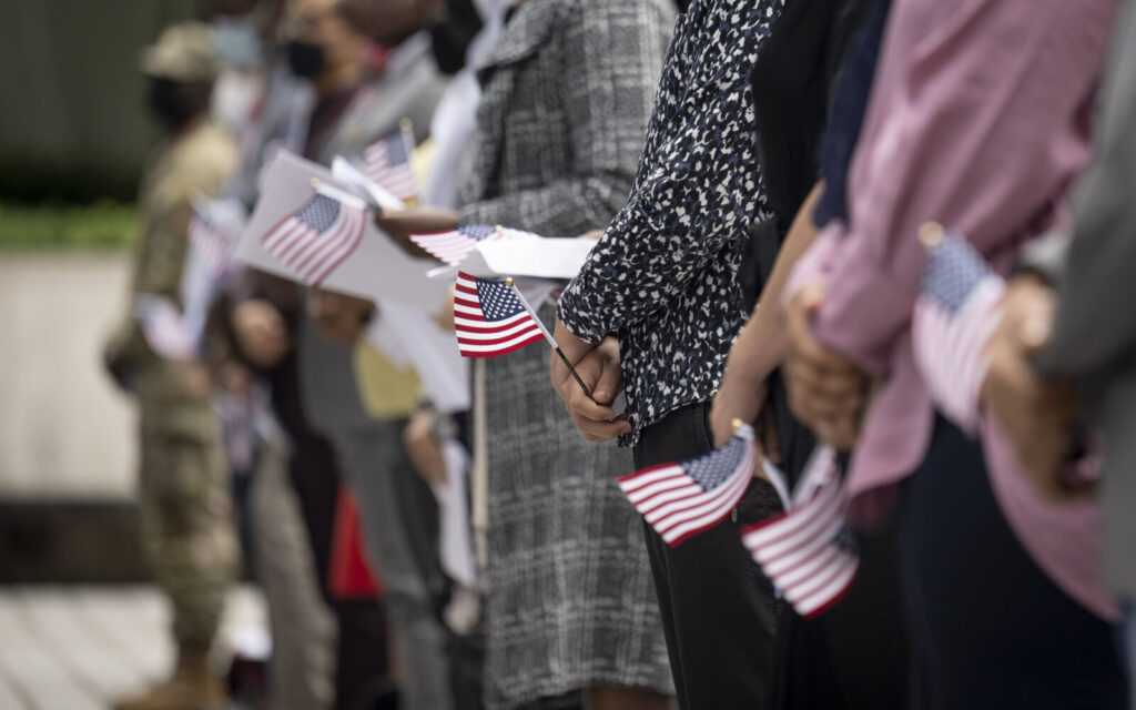 A foto mostra pessoas alinhadas seguram pequenas bandeiras dos Estados Unidos, em ambiente externo.