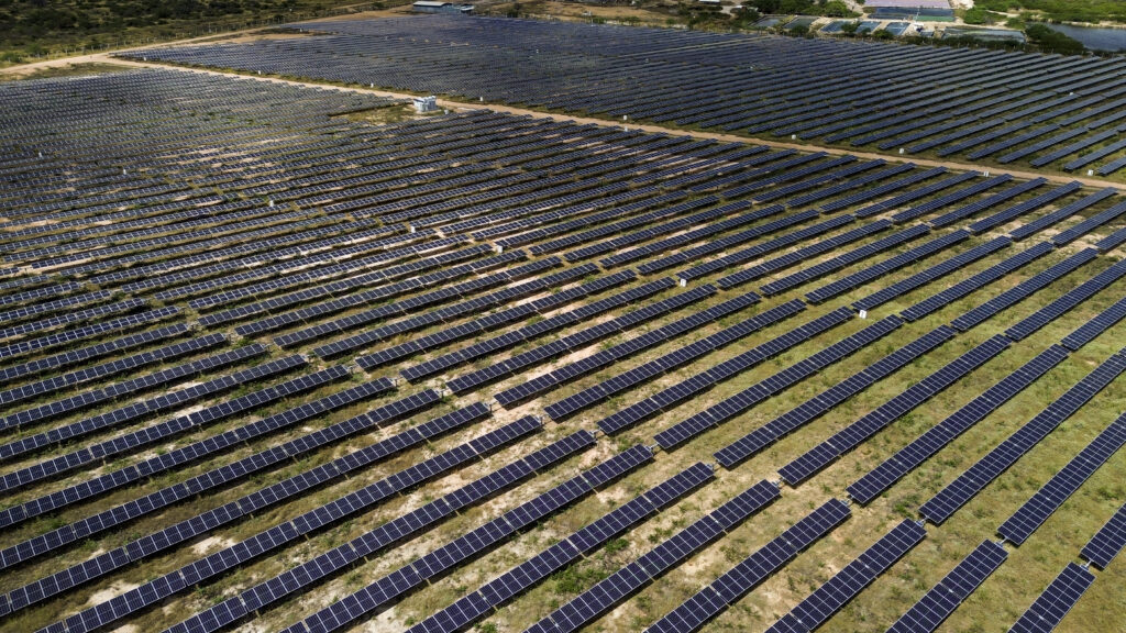 A imagem mostra extensa área ocupada por usinas solares – painéis fotovoltaicos dispostos em longas sequências formando fileiras paralelas que se estendem até o horizonte. Estão organizados em linhas retas, perfeitamente alinhadas, sugerindo uma instalação industrial de larga escala, com vista aérea levemente inclinada. Os painéis têm tons escuros (azulados ou acinzentados), refletindo sutilmente o céu. O solo entre e ao redor dos módulos parece ser de terra clara ou vegetação esparsa, contrastando com os painéis instalados sobre ele. No conjunto, o foco está na geometria repetitiva das fileiras de painéis solares — contrastando com o ambiente natural em volta, mas sem elementos humanos visíveis ou construções além dos módulos fotovoltaicos.