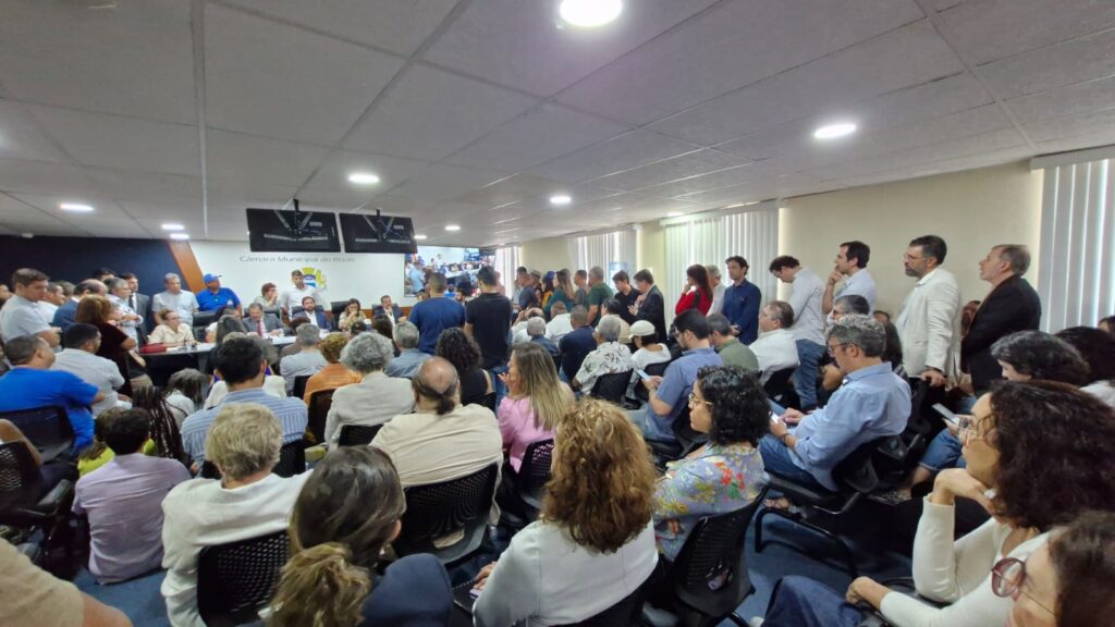 A foto mostra uma sala de reuniões da Câmara Municipal do Recife, bastante cheia de pessoas. Há homens e mulheres sentados em cadeiras e também em pé, ocupando todo o espaço disponível. No fundo, atrás de uma mesa grande, estão várias pessoas, aparentemente vereadores e autoridades, participando de uma sessão ou audiência pública. Acima deles, há dois monitores suspensos no teto, exibindo informações da sessão. A plateia é diversa, com pessoas de idades diferentes, algumas fazendo anotações, outras apenas assistindo com atenção. O ambiente transmite a ideia de um encontro formal, lotado e de grande interesse público.