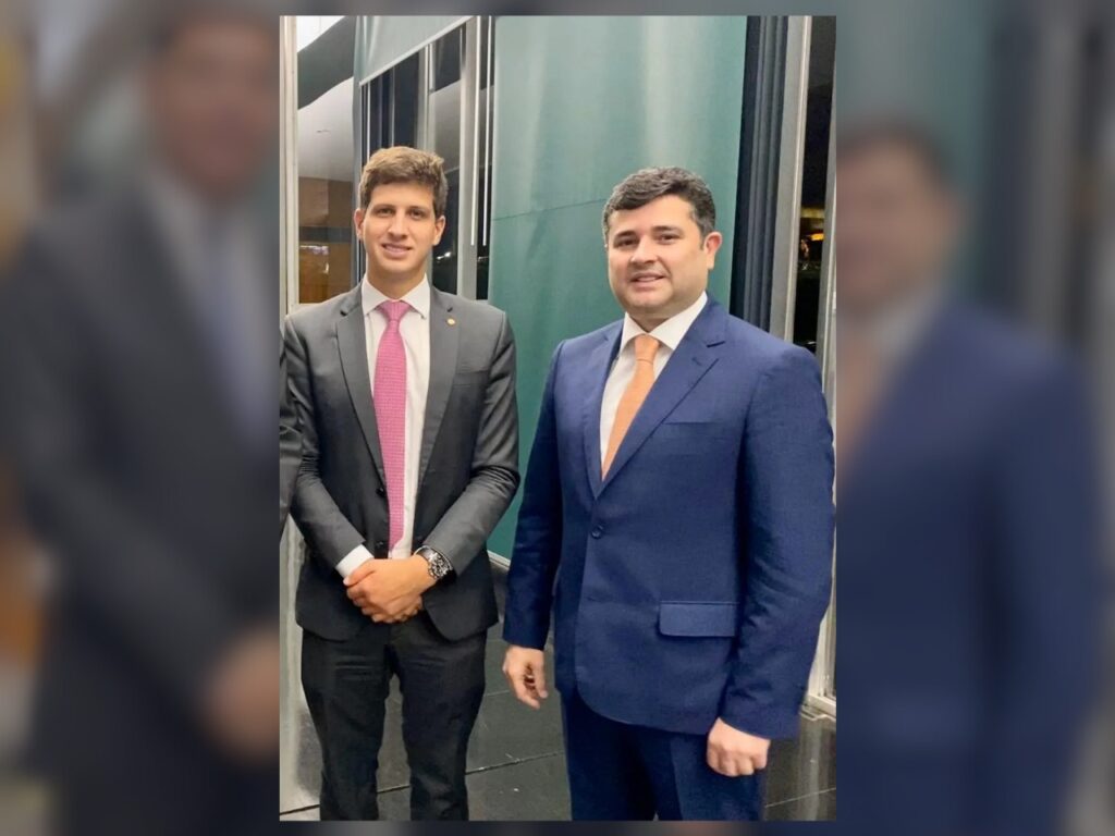 A foto mostra dois homens em pé, lado a lado, em um ambiente interno iluminado artificialmente, com paredes de vidro ao fundo. À esquerda está João Campos, homem jovem, de pele clara, cabelos castanhos curtos e penteados, usando terno escuro, camisa branca e gravata rosa. Ele sorri levemente e mantém as mãos unidas à frente do corpo. À direita está Eduardo da Fonte, homem de pele clara, cabelos curtos e escuros, com o rosto mais arredondado. Ele veste terno azul, camisa branca e gravata laranja, sorri discretamente e mantém o braço direito levemente à frente. Ambos aparecem em postura formal, transmitindo elegância e sobriedade.