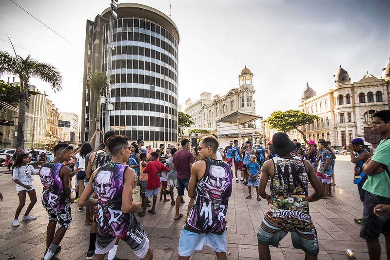 A imagem mostra um grupo de jovens dançando passinho no Marco Zero do Recife. Eles estão vestindo roupas coloridas, de costas, voltados para uma plateia que os observa.