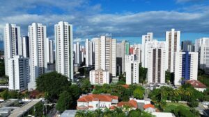 A imagem mostra a paisagem urbana da zona norte do Recife durante o final do dia, com uma bela luz dourada do pôr do sol iluminando o cenário. No primeiro plano, há uma rua ampla e tranquila, com algumas árvores ao lado. À esquerda da foto, é possível ver um edifício moderno de vários andares, com grandes janelas de vidro. À direita, há uma área verde com arbustos e mais árvores. Ao fundo, o céu está tingido de tons quentes, como laranja e amarelo, enquanto as nuvens refletem a luz do sol poente. A luz suave e dourada da tarde cria sombras longas nas construções e na rua.