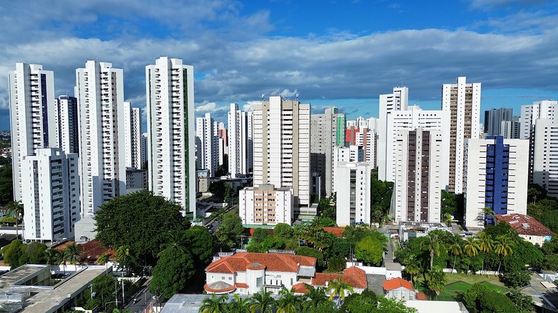 A imagem mostra a paisagem urbana da zona norte do Recife durante o final do dia, com uma bela luz dourada do pôr do sol iluminando o cenário. No primeiro plano, há uma rua ampla e tranquila, com algumas árvores ao lado. À esquerda da foto, é possível ver um edifício moderno de vários andares, com grandes janelas de vidro. À direita, há uma área verde com arbustos e mais árvores. Ao fundo, o céu está tingido de tons quentes, como laranja e amarelo, enquanto as nuvens refletem a luz do sol poente. A luz suave e dourada da tarde cria sombras longas nas construções e na rua.