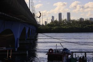 Na imagem, vemos um jovem saltando de uma ponte em direção ao rio, na Bacia do Pina, no Recife. O corpo dele está inclinado para trás, com os braços erguidos e as pernas dobradas, no momento exato em que está no ar. A ponte, pintada de azul em sua parte inferior, ocupa o lado esquerdo da foto. À frente, a água reflete a luz do céu, que está parcialmente nublado com nuvens claras. No fundo, aparecem prédios altos da cidade, atrás de uma faixa de árvores verdes. Na parte inferior direita da imagem, há um pequeno barco de pesca com boias e redes penduradas.